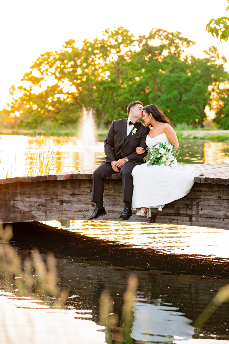 bride and groom sitting on bridge and kissing with sunset in the background of their sacramento wedding venue